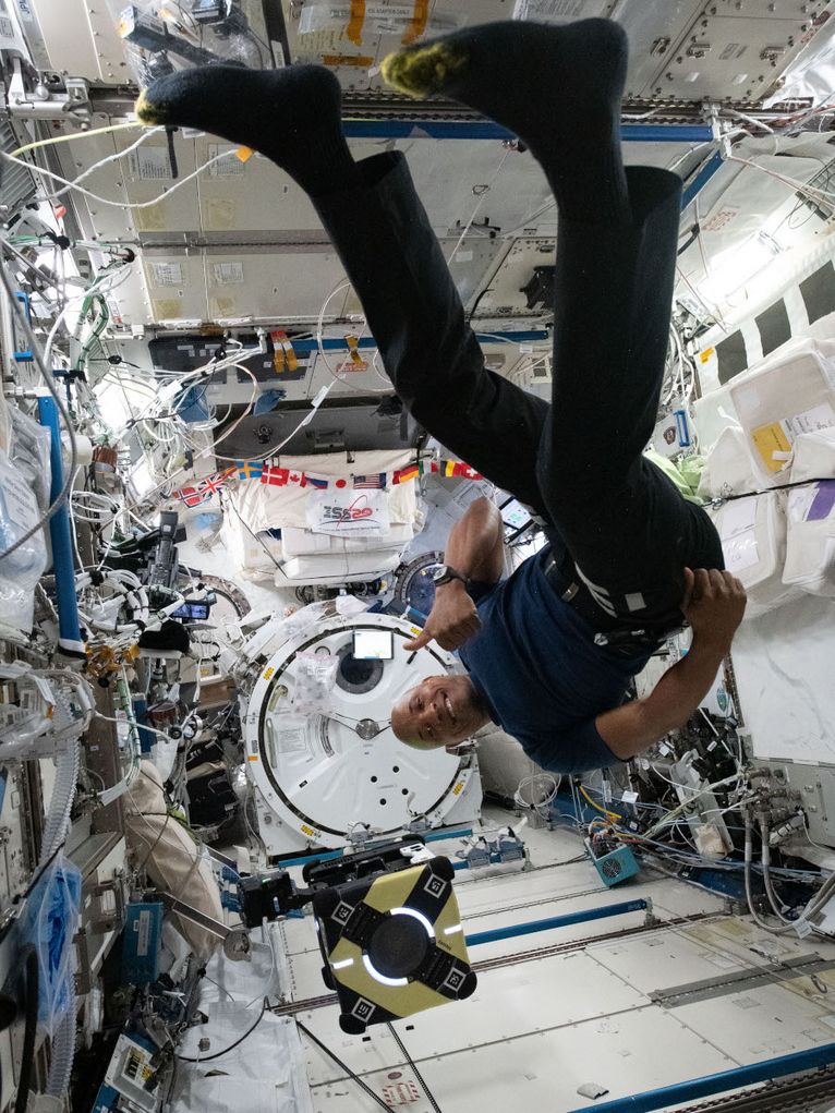 Person schwebt Kopfüber in einer Weltraumkapsel der ISS mit vielen Kabeln und dem Gecko-Roboter.