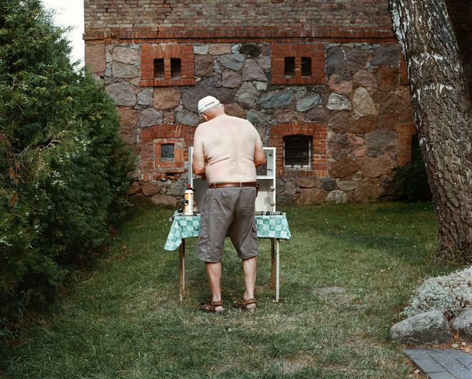 Ein älterer Mann mit freiem Oberkörper an einem Gartengrill, man sieht ihn von hinten.