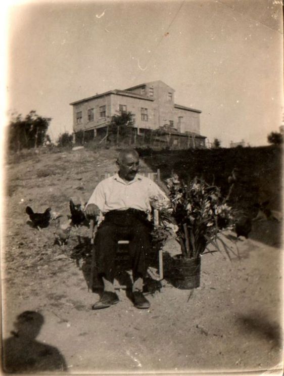 Ein Mann in einem Gartenstuhl vor einem erhöhten Haus. Neben ihm eine Vase mit Blumen, hinter ihm Hühner.