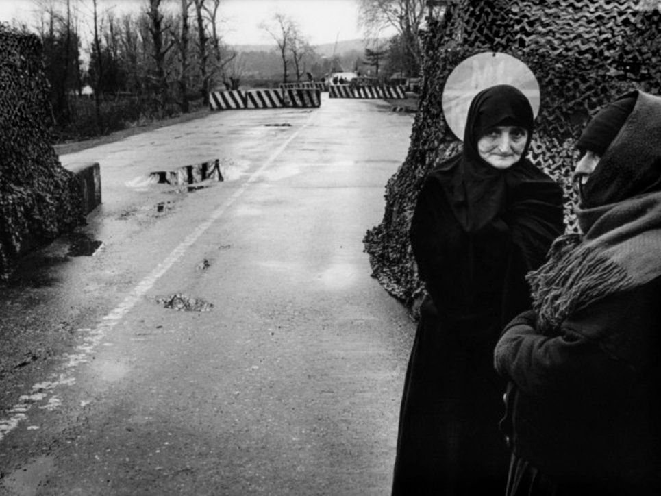 Schwarzweißfoto von zwei warm gekleideten Frauen am Straßenrand. Im Hintergrund Absperrungen und Wald.