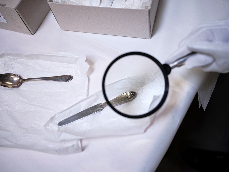 Unter einer Lupe ist die Gravur auf dem Griff eines Silbermessers zu sehen, auf dem Tisch liegt auch ein kleiner silberner Löffel.