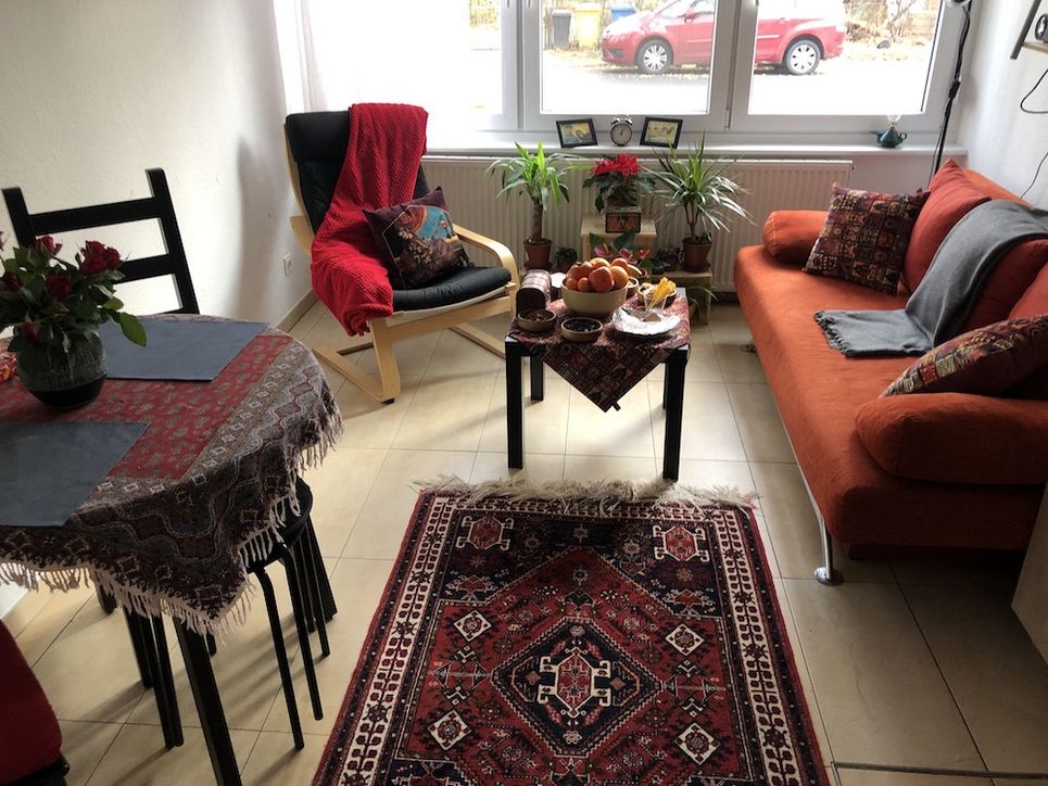 Gemütlich eingerichtetes Wohnzimmer, mit einem bunten Teppich, Sofa und Sessel, einem Tisch mit Blumen und einem kleinen Tischchen mit einer Obstschale. 