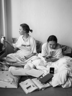 Zwei Mädchen sitzen mit Laptop, Handy und Schulbüchern im Bett.