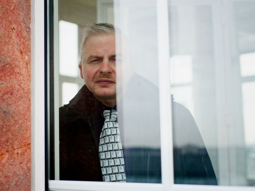 Jörg Muskatewitz blickt hinter einem Vorhang hervor aus einem Fenster.