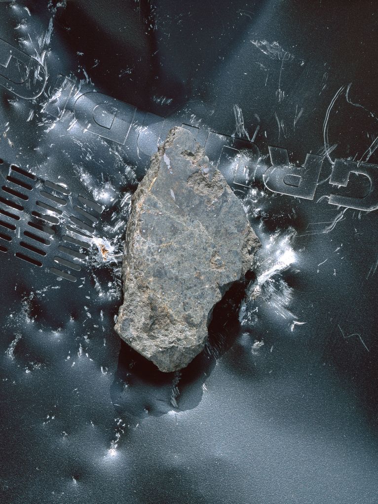 Grauer Meteorit, der auf das Metallgehäuse ein Geräts der Marke Grundig aufgetroffen ist und deutliche Spuren hinterlassen hat. 
