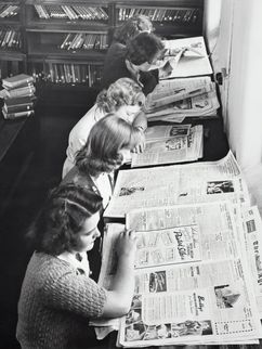 Junge Frauen sitzen in einer Reihe an einem Tisch und lesen Zeitung in einer Bibliothek