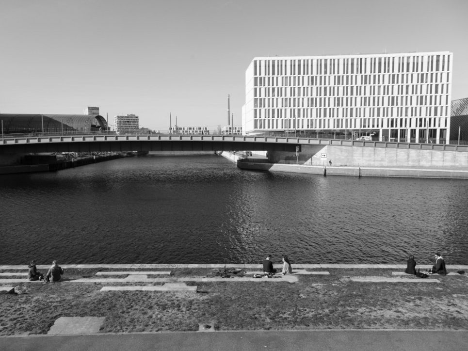 Menschen sitzen in Zweiergrüppchen am Wasser in einer Stadt.