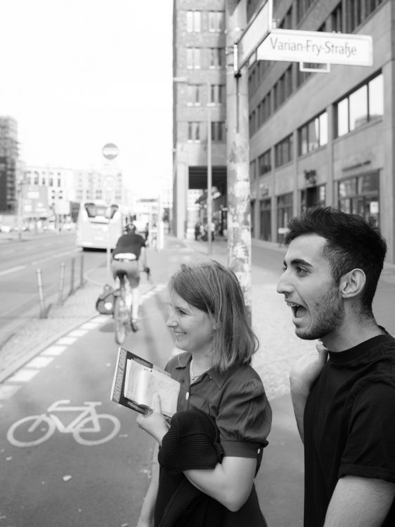Schwarzweißfoto von Ahmad Khalil und unserer Autorin Susanne Hörr auf einem Spaziergang durch Berlin-Neukölln. Im Hintergrund ein Straßenschild, Hochhäuser, ein Bus und ein Radfahrer.