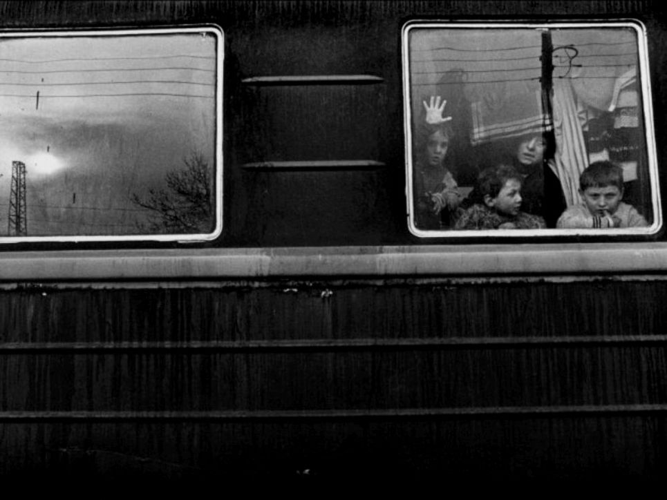 Ein Schwarzweißfoto. Durch das Fenster eines Zugwagons sind drei Kinder und eine Frau zu sehen. Masten und Stromleitungen spiegeln sich in den Fenstern.