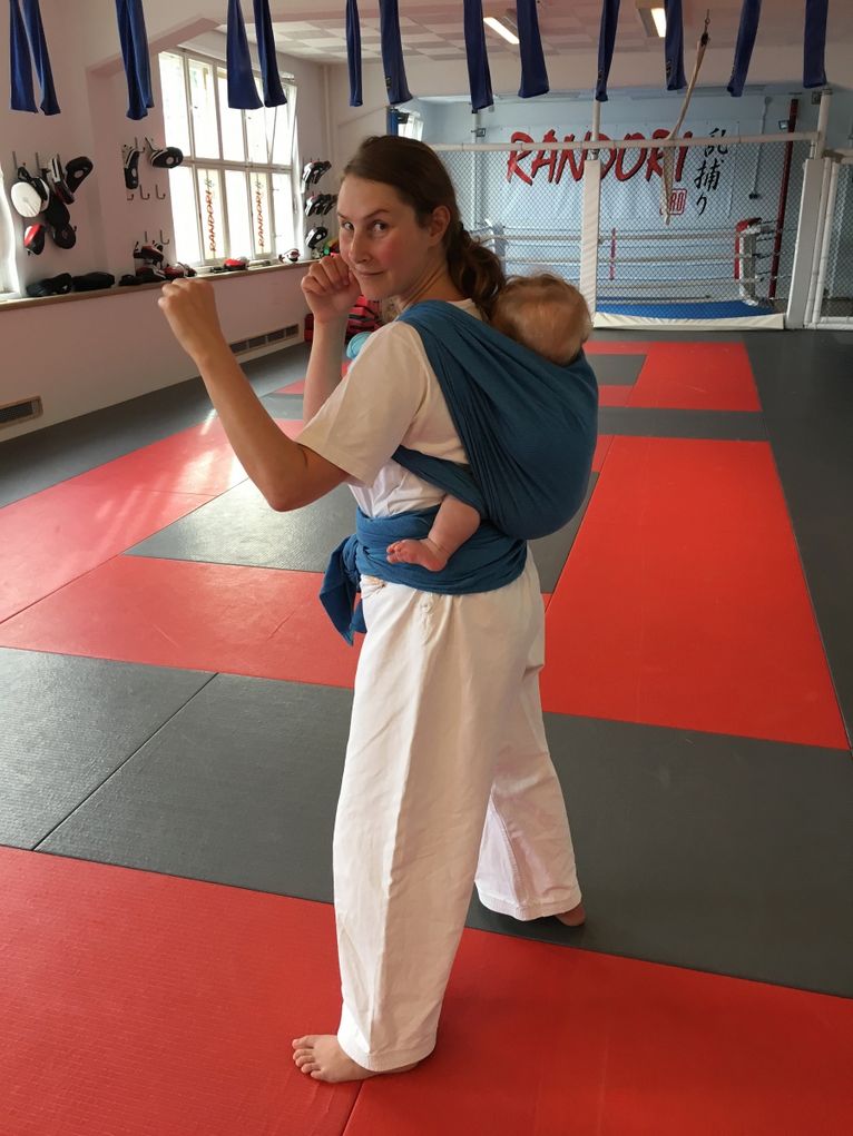 Mara Osswald in Jiu-Jitsu-Pose und weißem Anzug mit einem kleinen Kind im Tragetuch auf dem Rücken.