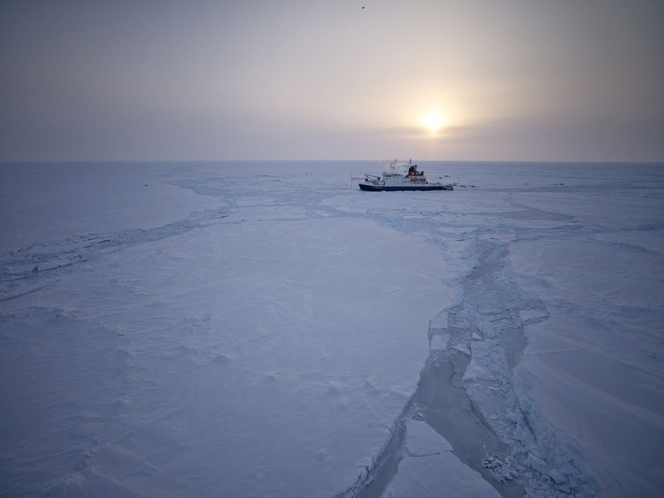 Das Forschungsschiff in der Ferne inmitten des Eismeers, fahle Sonne im Hintergrund.