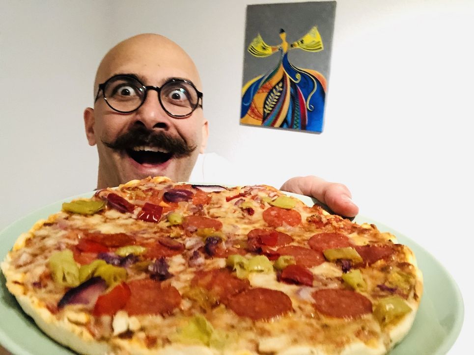 Nader Marzban hält lachend einen Teller mit einer Fertigpizza in die Kamera