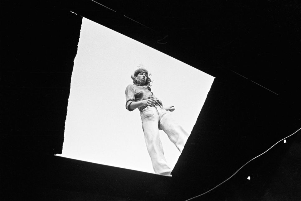 Schwarzweißfotografie. Ein Mann mit langen Haaren und weiter Hose von unten durch eine viereckige Öffnung fotografiert.