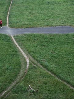 Von oben fotografierte, sich kreuzende Wege auf einer grünen Wiese, die in fünf Richtungen zeigen. Zwei Personen stehen an der Kreuzung.