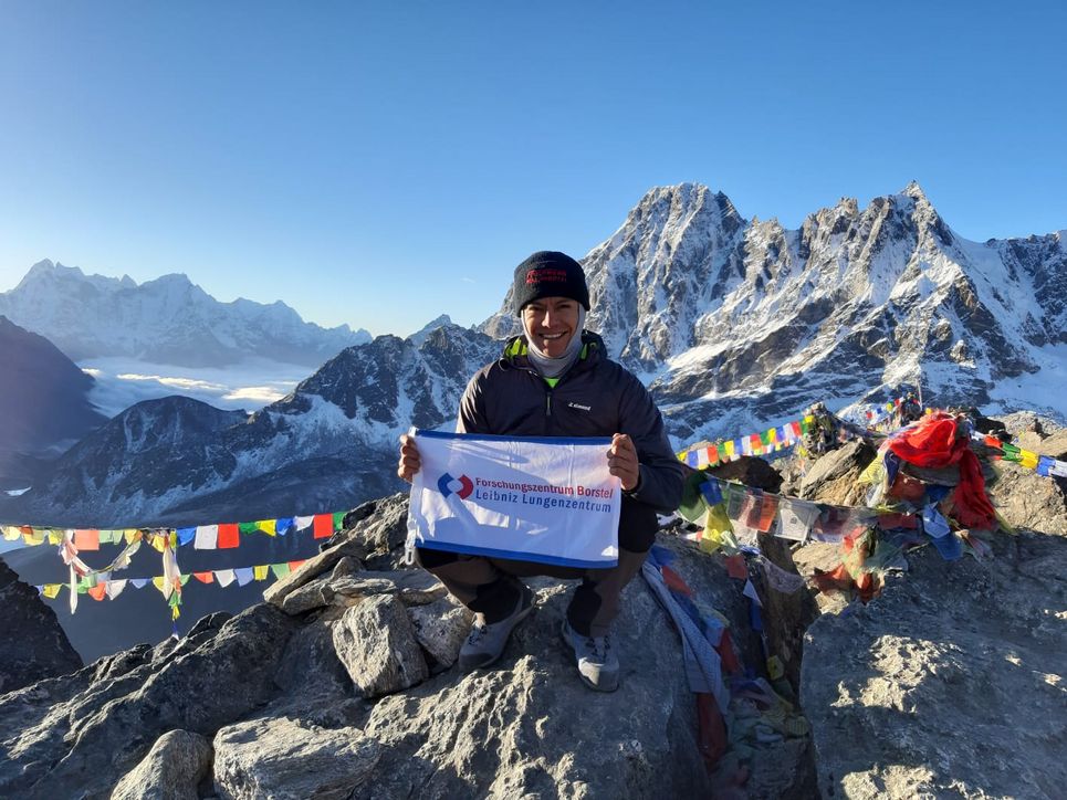 Bergsteigerin mit Banner des Forschungszentrum Borstel und Tibetfähnchen auf einem Gipfel.