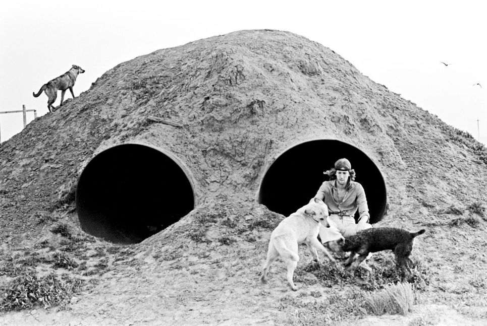 Schwarzweißfotografie. Der Mann mit zwei Hunden bei einem Hügel, aus dem zwei große Rohre ragen.