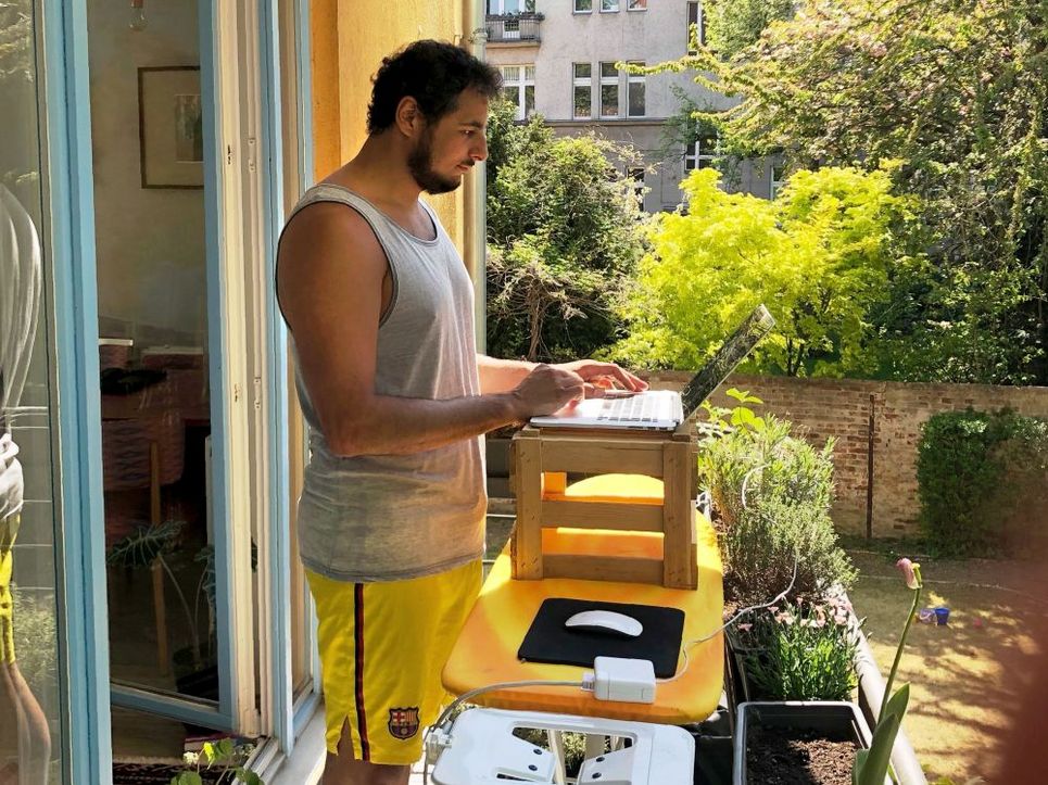 Ein junger Mann steht auf einem bepflanzten Balkon und arbeitet am Laptop, der auf einer Holzkiste auf einem Bügelbrett steht. 