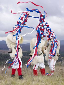 Drei Personen auf einer Anhöhe. Sie sind in Pelze gekleidet, tragen Kuhglocken um die Hüfte und haben riesige blau-rot-weiße Hüte mit Bändern auf dem Kopf.