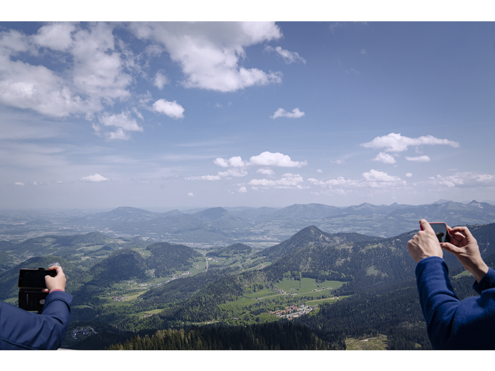 Großes Bergpanorma vom Obersalzberg. An beiden Seiten ragen Arme von Menschen ins Bild die Fotos von der Aussicht machen. Leibniz Magazin 