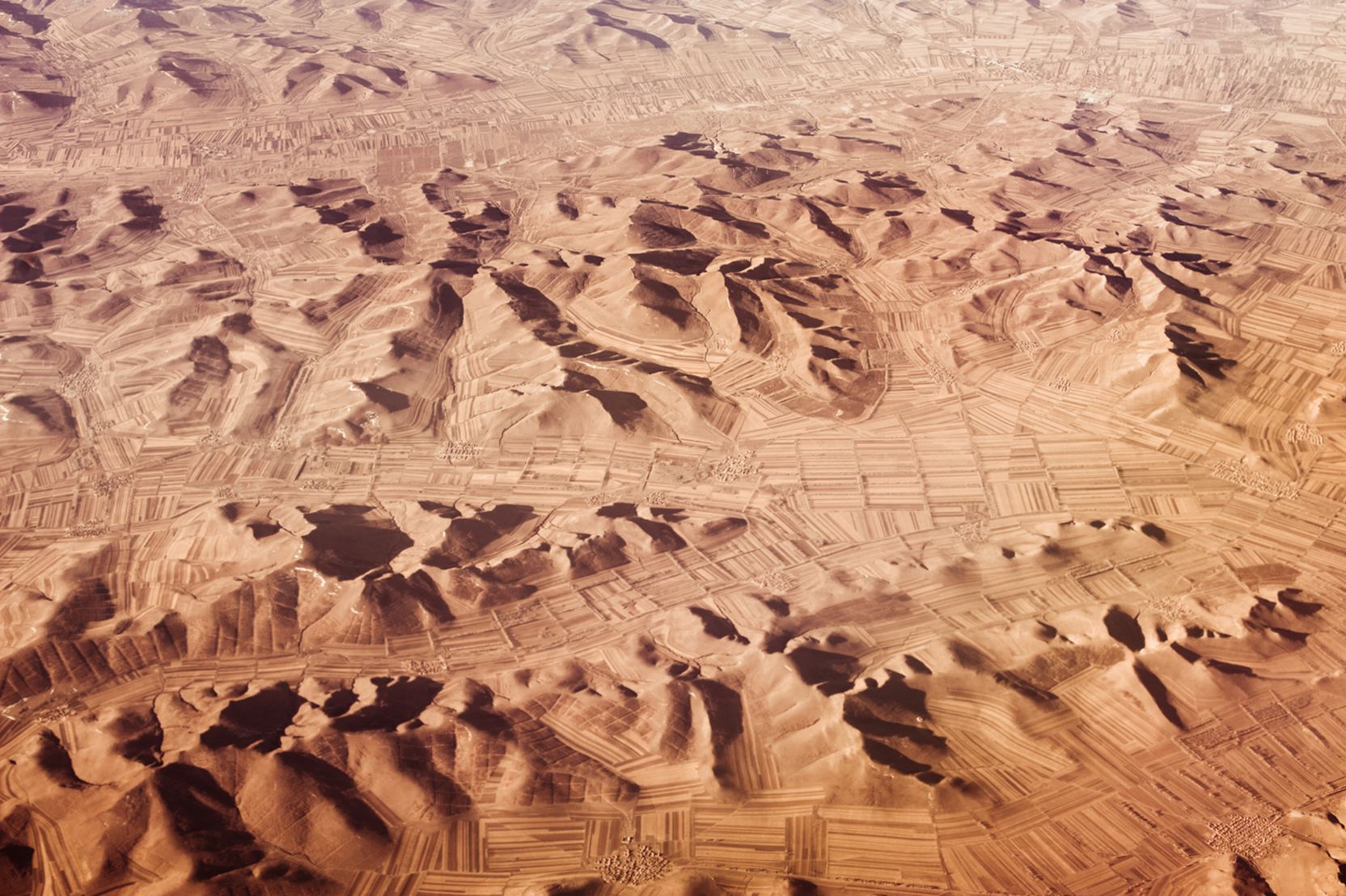 Luftbildaufnahme einer sandigen, bergigen Landschaft mit Strukturen, die Felder und Dörfer vermuten lassen