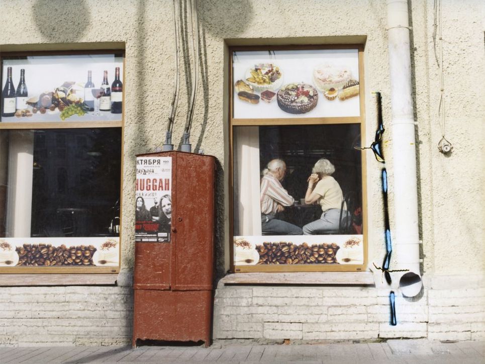 Durch das Fenster eines Osteuropäischen Lokals sieht man ein älteres Ehepaar an einem Tisch sitzen. Die Fenster des Lokals sind mit Fotos von Wein, Desserts und Kaffeebohnen dekoriert. Ein rostbrauner Stromkasten steht zwischen den Fenstern. 