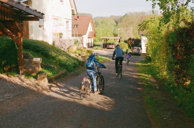 Eine erwachsene Person und zwei Kinder von hinten auf ihren Fahrrädern in einem Wohngebiet.