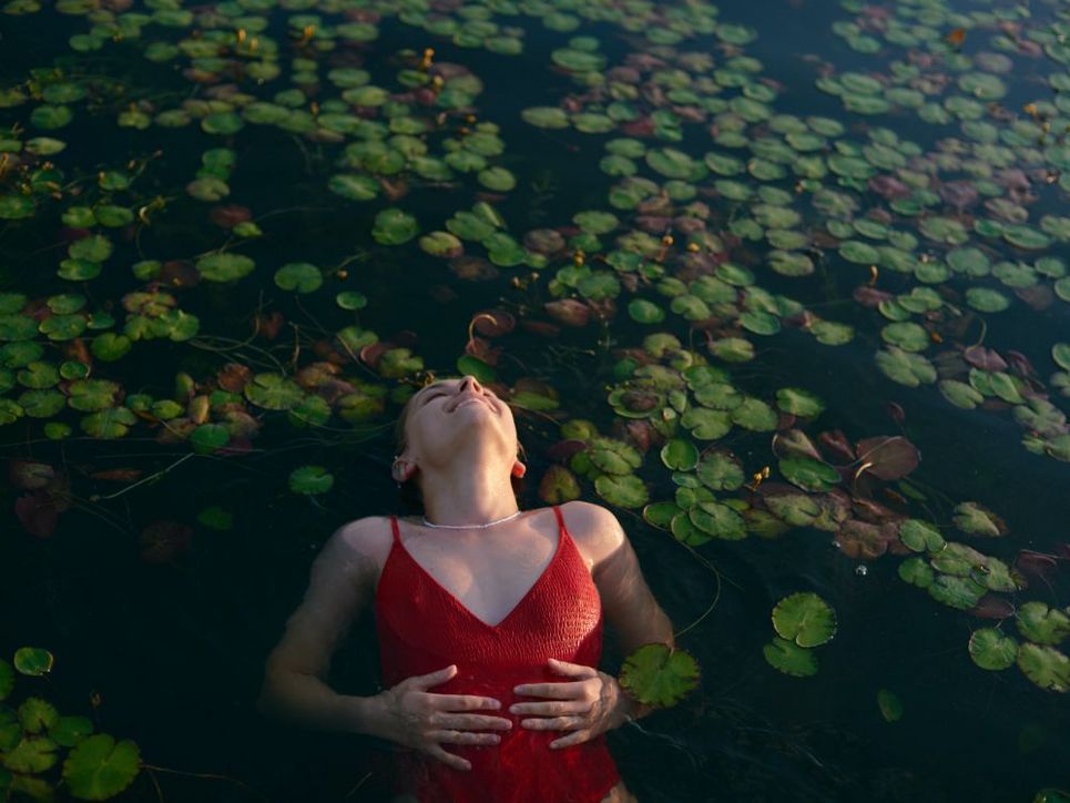 Eine Frau in einem roten Badeanzug liegt in Rückenlage im Wasser. Hinter ihr schwimmen Seerosen.