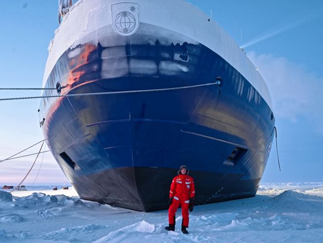 Martin Radenz im roten Schneeanzug steht vor dem großen, dunklen Bug des Schiffes. Im Verhältnis sieht er sehr klein aus.