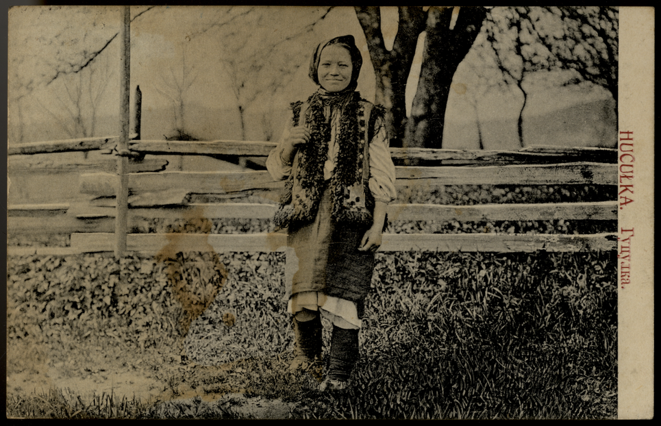 Postkarte, die ein bäuerlich gekleidetes Mädchen zeigt, das vor einem Lattenzaun steht.