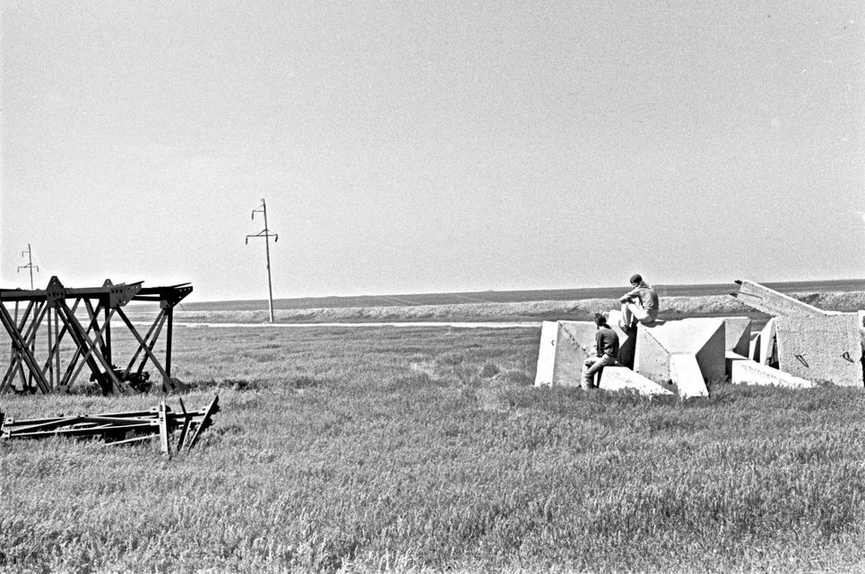 Zwei Personen sitzen auf Bauteilen in einer weiten Graslandschaft. Schwarzweiß-Fotografie.