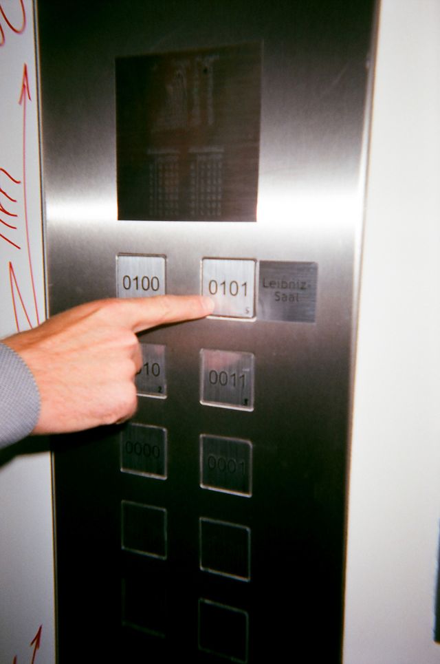 Ein Finger berührt die Taste eines Aufzugs, die mit »Leibniz-Saal« beschriftet ist und die Nummer 0101 trägt.