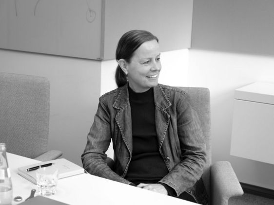 Bettina Hörstrup sitzt an einem Tisch und hat ihren Kopf im Gespräch zum rechten Bildrand gedreht. Foto in schwarz-weiß