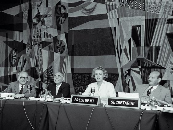 Maria Daelen an einem Konferenztisch, vor ihr ein Schild mit der Aufschrift »President«. Rechts zwei Herren, links ein weiterer. 