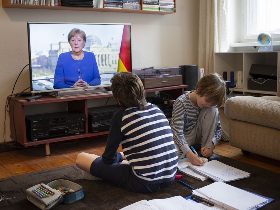 Zwei Jungen sitzen mit Schulheften auf dem Fußboden vor dem laufenden Fernseher, Angela Merkel zeigt.
