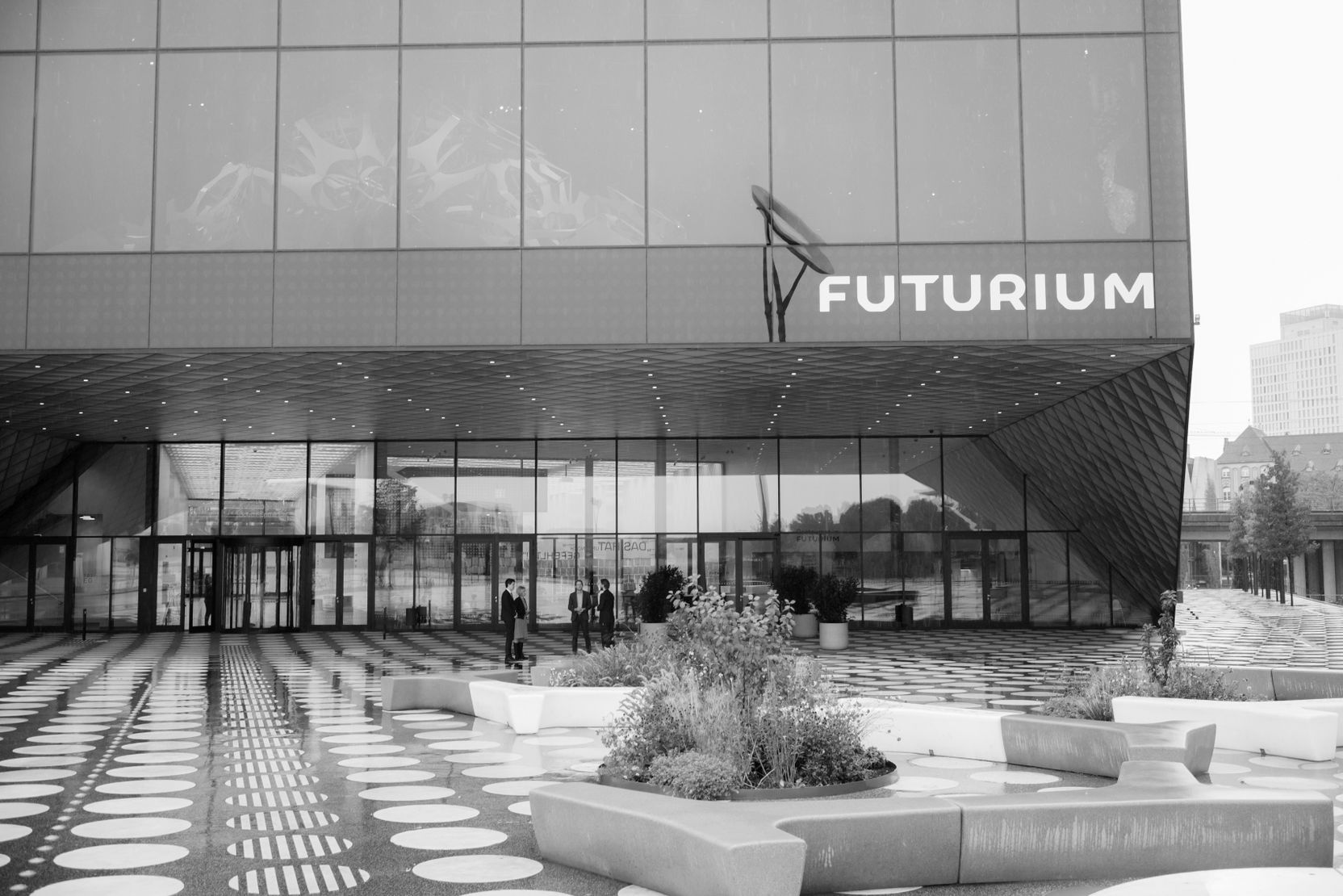 Schwarzweißfoto. Sunhild Kleingärtner, Stefan Brandt, Swen Schulz und unser Moderator Frank Steinhofer unterhalten sich vor dem verglasten Eingangsbereich des Futuriums in Berlin.