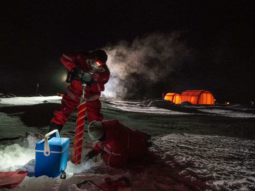 Eine Person bohrt einen großen, dicken Bohrer in die Eisfläche, eine weitere liegt am Boden. Im Hintergrund leuchtende Zelte.