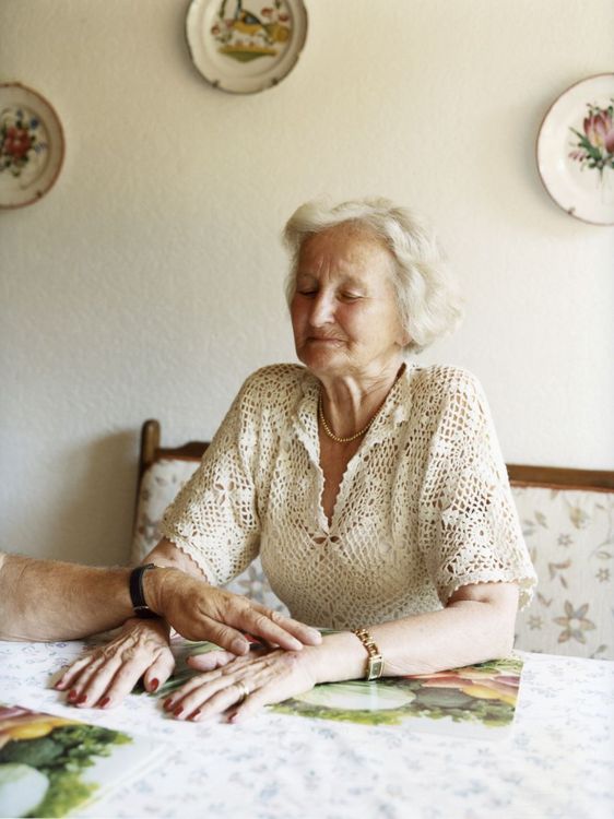 Eine ältere Frau sitzt auf einer Küchenbank, ihre Arme hat sie vor sich auf den Tisch gelegt. Eine andere Person legt seine Hand über ihre Hände.