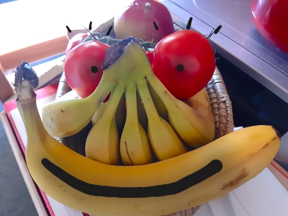 Obst-Berg mit Bananen, Tomaten und einem Äpfeln ist mit Augen und einem Mund bemalt und sieht aus wie ein Gesicht.