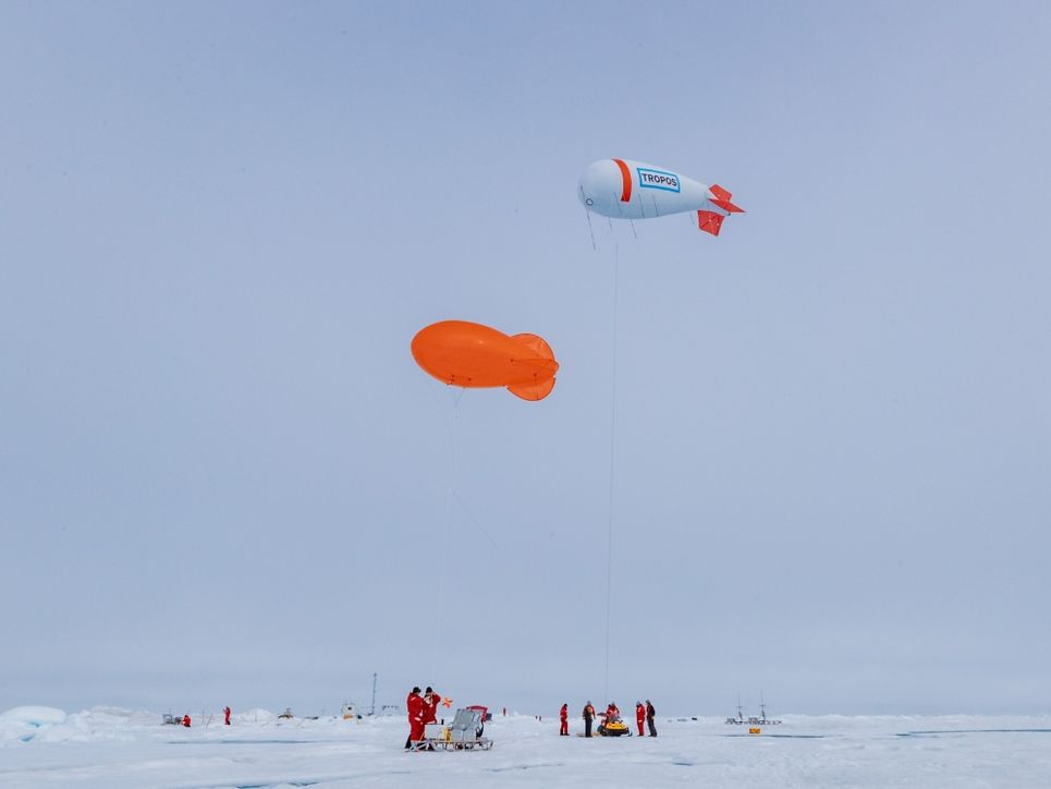 Ein orangener und ein weiß-rot-blauer Fesselballon in der Luft, mehrere Personen und Gerätschaften auf der Eisfläche.