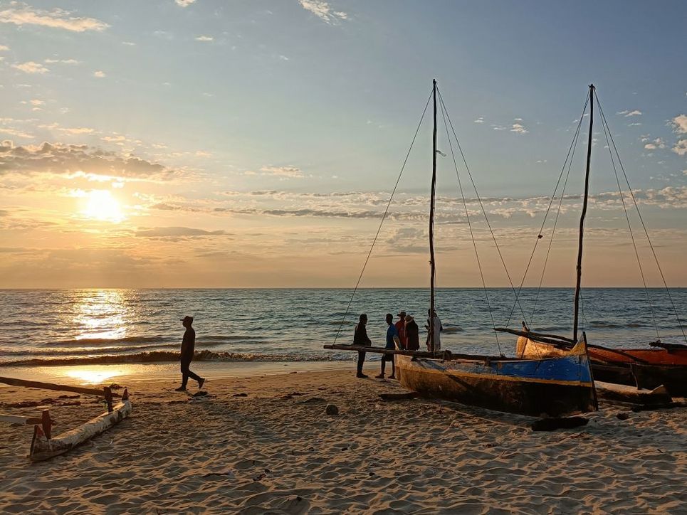 Strand bei Sonnenuntergang mit Piroge, einem langen hölzernen Boot und vereinzelten Personen.