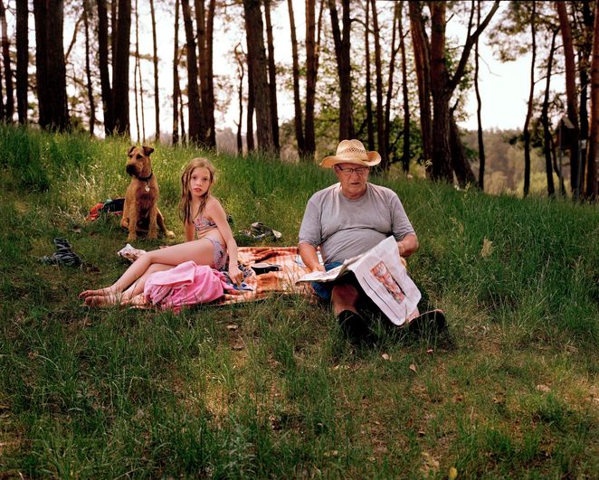 Ein älterer Mann, der Zeitung liest und ein Mädchen im Bikini auf einer Picknickdecke im Wald, hinter ihnen ein Hund.