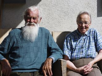 Zwei ältere Männer auf Holzstühlen vor einer sonnigen Hauswand.