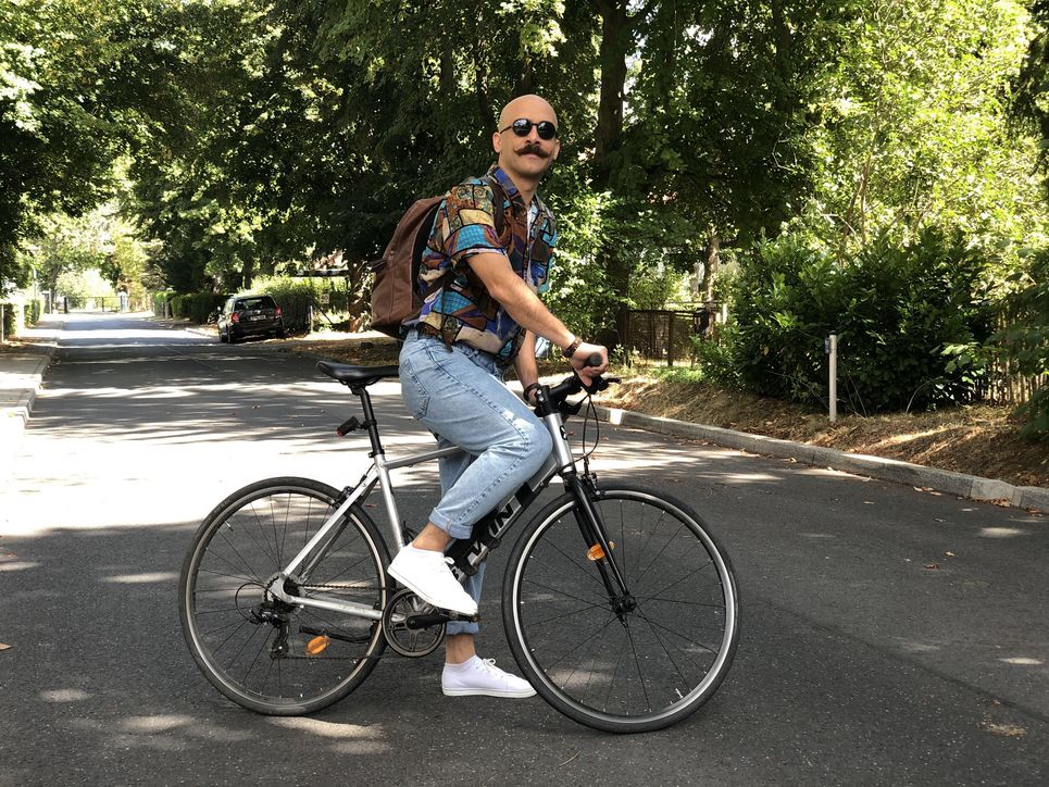 Nader Marzban steigt auf´s Fahrrad, mit Sonnenbrille, Rucksack, Jeans und buntem Hemd.