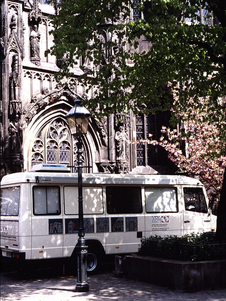 Ein weißer Bus, das Labormobil, parkt vor einer Kirche unter einem Baum.