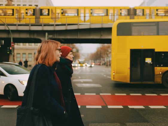 Annalisa Martin und Svenja Beller überqueren die Straße, neben ihnen ein gelber Doppeldecker-Bus, ein Abbiegendes Auto und hinter ihnen die hochgelegte U-Bahn.