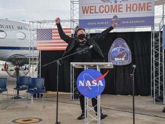Auf einer Bühne feiert Elon Musk die Rückkehr der SpaceX-Astronauten Dough Hurley und Bob Behnke. Im Vordergrund das Logo der NASA, im Hintergrund die amerikanische Flagge und ein Flugzeug.