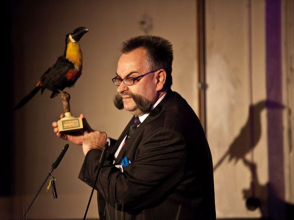 Johannes Vogel mit einem ausgestopften Papageien in der Hand auf einer Bühne mit Mikrofonen.
