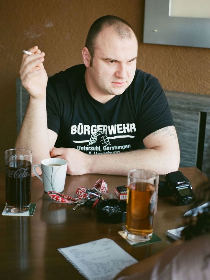Andreas Niebling mit Zigarette und Getränken. Auf seinem schwarzen T-Shirt steht: Bürgerwehr.