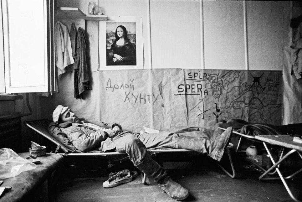 Schwarzweißfotografie eines Mannes in Arbeitskleidung. Er liegt auf einem Feldbett, über ihm das Bild der Mona Lisa, aufgehängte Kleidung und eine, zum Teil mit kyrillischen Buchstaben beschriftete Wand.
