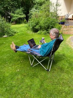 Ein Mann im Campingstuhl im Garten, mit Laptop, Getränk und Headset.
